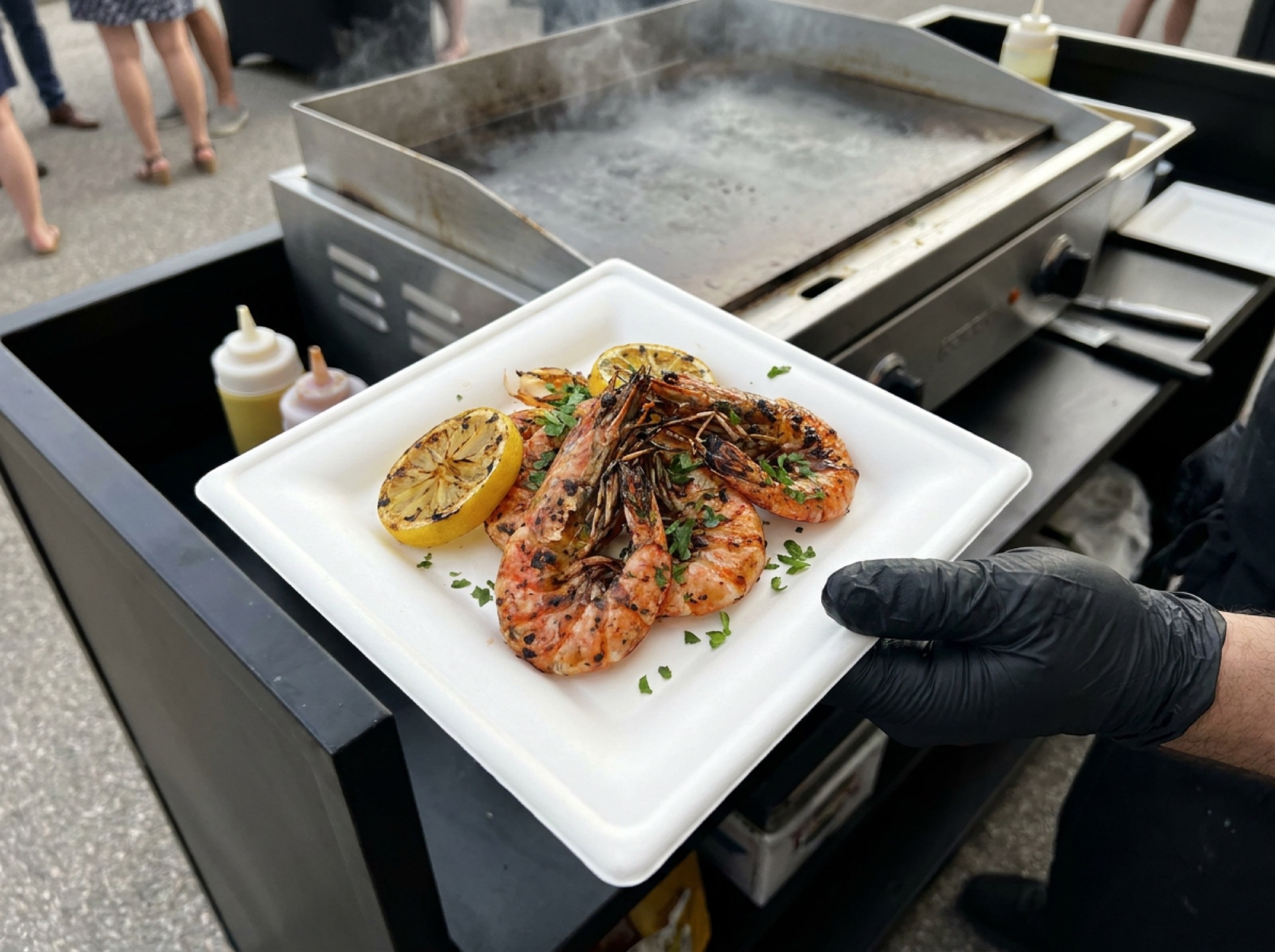 Gamba's a la plancha op wit suikerrietbord naast de hete plancha, vastgehouden met zwarte handschoenen